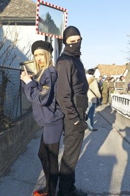 Eine Frau und ein Mann in Polizeiuniformen stehen auf einer Straße, wobei die Frau einen Pokal hält und der Mann eine Maske trägt. Hinter ihnen versammeln sich Menschen, und ein Gebäude ist im Hintergrund zu sehen.