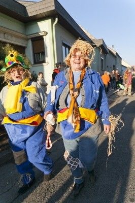 Zwei Menschen in Strohkostümen gehen während einer Parade die Straße entlang, mit Gebäuden und Menschen im Hintergrund.