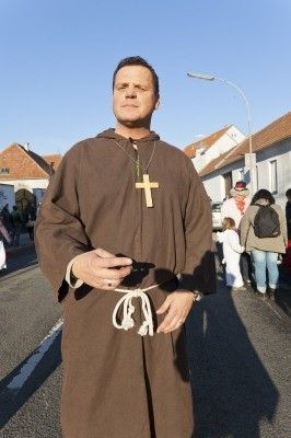 Ein Mann, der als Mönch verkleidet ist, steht auf einer Straße, trägt eine braune Kutte und ein Kreuzhalsband, mit Gebäuden und Menschen im Hintergrund.