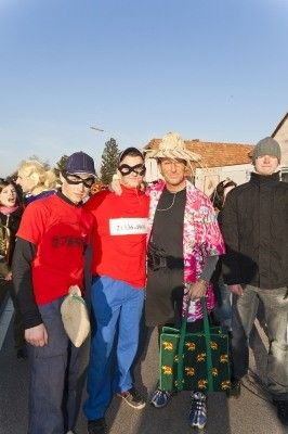 Vier Menschen in Kostümen stehen auf einer Straße. Zwei Männer tragen rote Hemden, einer hat eine Maske. Eine Frau in einem Blumenkleid hält eine grüne Tasche. Ein Mann in einem Hut steht hinter ihnen.