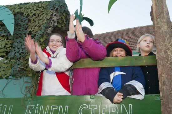 Vier Kinder in Kostümen stehen in einem geschmückten Wagen, winken und lächeln in die Kamera. Der Wagen hat einen grünen Hintergrund mit deutschem Text.