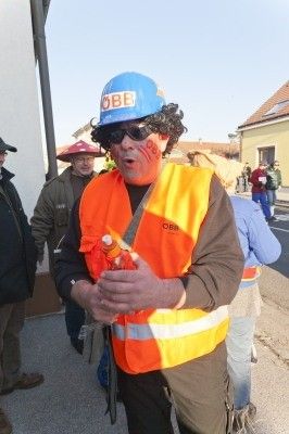 Ein Mann in einer Bergmanns-Kostüm mit einem blauen Helm und oranger Weste, eine Flasche haltend, steht auf einer Straße mit anderen Leuten im Hintergrund.