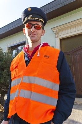 Ein Mann in einer reflektierenden orangefarbenen Weste mit 'OBB' darauf steht vor einem Gebäude, trägt eine Sonnenbrille und einen Hut.