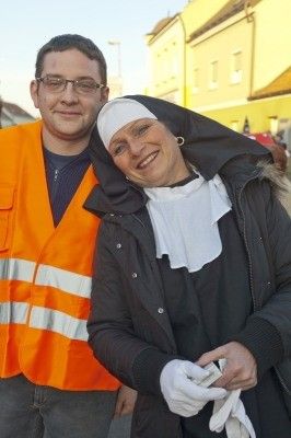 Ein Mann in einer orangefarbenen Sicherheitsweste und eine Frau, die als Nonne gekleidet ist, stehen lächelnd für ein Foto auf einer Stadtstraße.