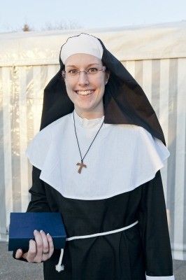 Eine lächelnde Nonne in einem schwarzen und weißen Habit, mit Brille und Kreuzkette, hält ein blaues Buch.