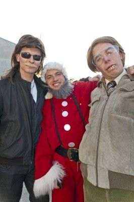 Drei Männer posieren für ein Foto; einer in einem Weihnachtsmannkostüm, einer in einer Jacke mit Schnurrbart und einer in einer Brille und Krawatte.