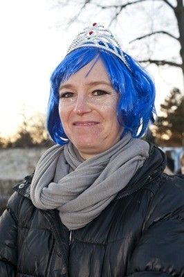 Eine Frau mit blauen Haaren, die eine Krone und einen grauen Schal trägt, lächelt vor einem Baum.