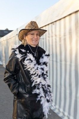 Eine Frau in einer schwarzen Lederjacke und einem Leopardenmuster-Hut steht draußen, lächelt und trägt eine weiße Federboa.