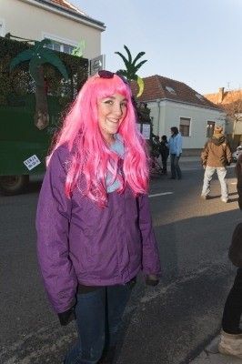 Eine Frau mit rosa Haaren und einer rosa Perücke steht am Straßenrand und lächelt in die Kamera. Hinter ihr ist ein Wagen mit Palmen und einem grünen Lastwagen zu sehen.