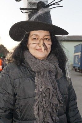 Eine Frau verkleidet als Hexe, trägt einen schwarzen Hut mit Spinnennetzen, einen grauen Schal und Make-up im Gesicht, steht draußen.