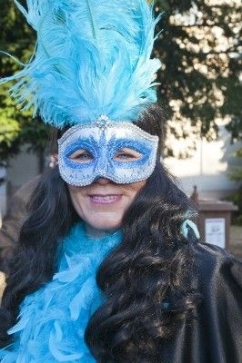 Eine Frau mit langen schwarzen Haaren, die eine blaue Federhaube und eine blaue Maske trägt, lächelt und steht im Freien.