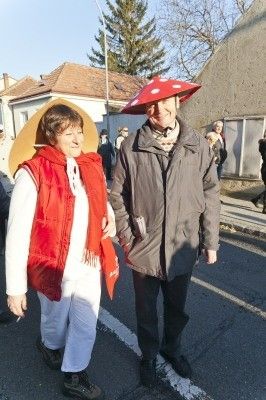 Ein Mann und eine Frau in Kostümen gehen auf einer Straße. Die Frau trägt einen roten Pilzhut und eine rote Weste. Der Mann trägt einen grauen Mantel. Sie halten sich an den Händen. Hinter ihnen sind Gebäude und Menschen, die auf der Straße gehen.