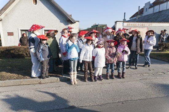 Eine Gruppe von Kindern und Erwachsenen in roten Pilzkappien steht in einer Reihe auf einer Straße. Hinter ihnen befinden sich Gebäude, eines mit einem Schild, das 'Arabergestut' besagt.
