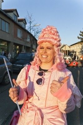 Eine Frau in einem rosa Kostüm mit Perücke und Sonnenbrille auf der Straße, hält eine Zauberstange und gibt einen Daumen hoch.