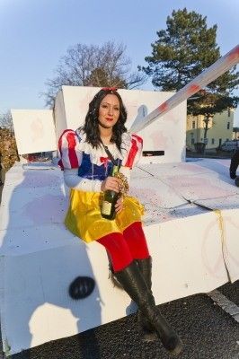 Eine Frau in einer Schneewittchen-Verkleidung sitzt auf einer weißen Plattform und hält eine Flasche Champagner in der Hand, mit einem großen weißen Objekt hinter ihr.
