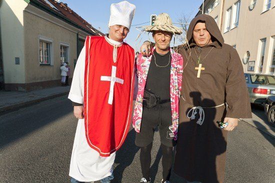 Drei Männer in Kostümen stehen auf einer Straße; einer trägt eine rote Robe mit einem Kreuz, ein anderer einen Strohhut und der dritte eine braune Robe mit einem Kreuz-Halsband.
