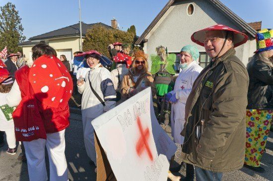 Eine Gruppe von Menschen in verschiedenen Kostümen, darunter Pilze und Feenflügel, versammelt sich um einen Mann, der ein Schild mit einem roten Kreuz in der Hand hält.