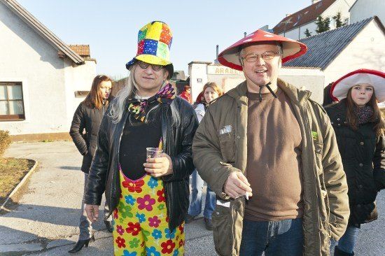 Zwei Männer in bunter Kleidung, einer trägt einen Hut, stehen zusammen auf einer Straße. Hinter ihnen geht eine Frau, und im Hintergrund befinden sich Gebäude und ein Schild.