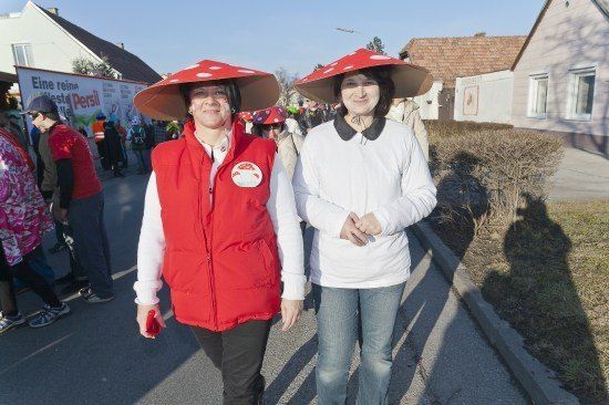 Zwei Frauen in Pilzhuten gehen eine Straße entlang, eine in einem roten Weste und die andere in einem weißen Hemd. Sie sind von anderen Menschen und einem Banner im Hintergrund umgeben.