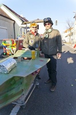 Zwei Personen in Militäruniformen stehen neben einem grünen Fahrzeug, möglicherweise einem Anhänger, auf einer Straße. Einer hält ein gelbes Objekt, und ein buntes Fahrzeug steht hinter ihnen.