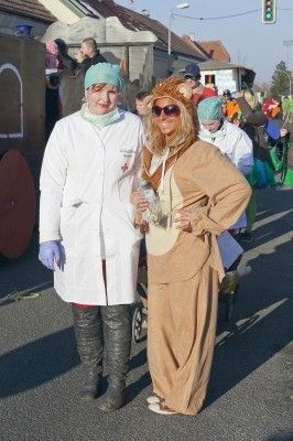 Zwei Personen in Kostümen, eine als Ärztin und die andere als Löwe, stehen auf einer Straße während einer Parade mit mehreren Personen im Hintergrund.