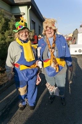Zwei Menschen in Strohkostümen, einer mit einem Sonnenblumenhut und der andere mit einem Schal, gehen die Straße entlang.