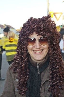 Eine Frau mit lockigem braunem Haar, Sonnenbrille und brauner Jacke lächelt im Freien.