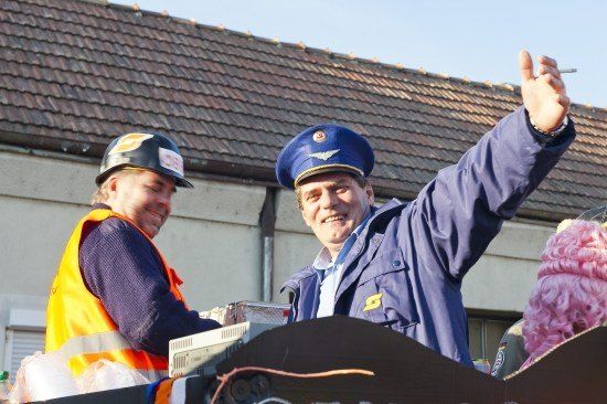 Zwei Männer in Kostümen, einer trägt einen Feuerwehrhelm und eine Weste, der andere ein Pilotenuniform, winken der Menge zu. Dahinter befindet sich ein Gebäude mit einem Ziegeldach.