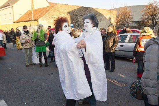 Zwei Personen in weißen Roben und mit bemalten Gesichtern tanzen auf einer Straße während eines Festivals, mit Zuschauern und geparkten Autos im Hintergrund.