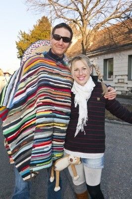 Ein Mann und eine Frau stehen draußen in Winterkleidung. Der Mann ist in eine bunte gehäkelte Decke gewickelt, und die Frau hat einen weißen Schal und hält ein Kuscheltier. Sie stehen vor einem Haus mit einem Baum.