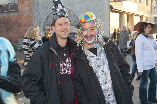 Zwei Männer in Kostümen, einer mit einem Zaubererhut und der andere mit einer Clownsmaske, stehen zusammen auf einer Straße. Hinter ihnen gehen mehrere Leute, und ein Gebäude mit Vordächern ist sichtbar.