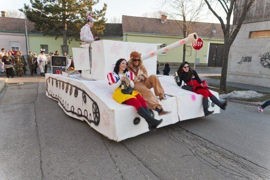 Vier Frauen in Kostümen sitzen auf einem dekorierten Panzerwagen in einem Umzug, mit einem Stoppschild und Gebäuden im Hintergrund.