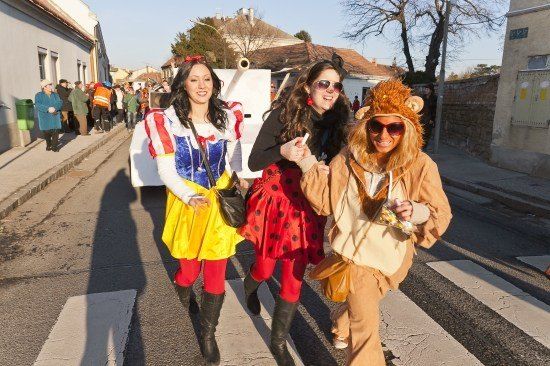 Drei Frauen in Kostümen überqueren eine Straße, mit einer Menschenmenge und einem Fahrzeug im Hintergrund.