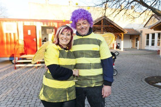 Ein Mann und eine Frau in Bienenkostümen stehen eng beieinander und lächeln in die Kamera. Hinter ihnen ist ein roter Container und ein Kinderwagen zu sehen.