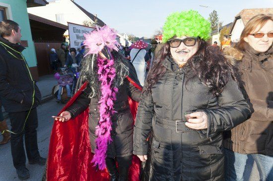 Zwei Frauen in Kostümen, eine mit einem rosa Federboa und die andere mit einer grünen Perücke, stehen auf einer Straße mit einer Menge Leute im Hintergrund.