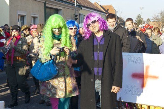 Eine Gruppe von Menschen in bunten Kostümen und Perücken steht auf einer Straße, einige spielen Musikinstrumente. Ein Mann mit lila Haaren und Sonnenbrille steht vor einem Whiteboard mit einem roten Kreuz.