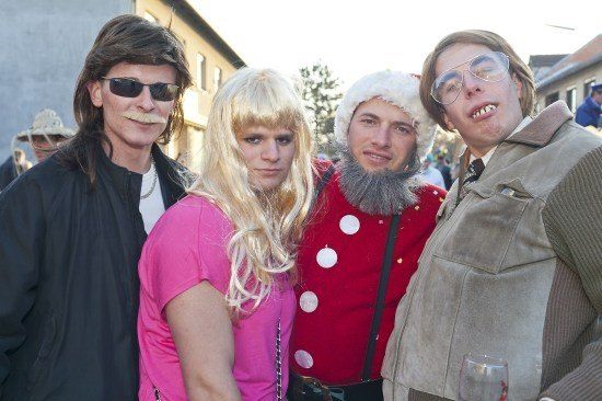 Vier Menschen in Kostümen, zwei Männer und zwei Frauen, posieren für ein Foto. Der Mann links hat lange Haare und einen Schnurrbart, die Frau neben ihm hat blonde Haare. Der Mann in der Mitte trägt einen weißen Hut und einen roten Mantel mit weißen Punkten. Der Mann rechts hat eine Brille und einen Bart.