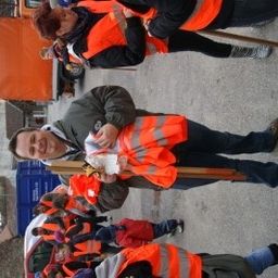 Eine Gruppe von Menschen in orangefarbenen Sicherheitswesten steht auf der Straße. Ein Mann hält einen Stock und lächelt.