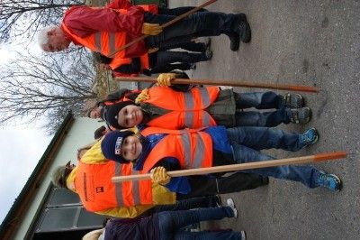 Eine Gruppe von Menschen, einschließlich Kindern, steht auf der Straße und trägt orange Westen. Sie halten Stöcke in den Händen. Sie scheinen an einer Gemeinschaftsaktion zur Müllentsorgung teilzunehmen.