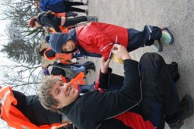 Ein Mann kniet und lächelt, während er einem Kind in einer roten Jacke hilft, einen Schuh anzuziehen. Sie sind von anderen Menschen in orangefarbenen Westen umgeben.