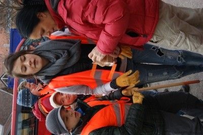 Eine Gruppe von Personen in Sicherheitswesten und Handschuhen, eine hält einen Stock, scheint gemeinsam an einer Aufgabe im Freien zu arbeiten.