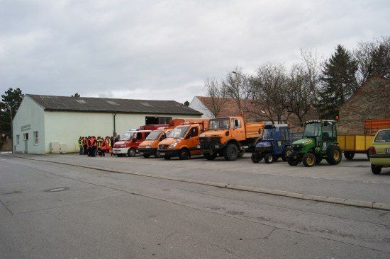 Mehrere Fahrzeuge, darunter Lastwagen und ein Traktor, sind vor einem Gebäude geparkt, in dessen Nähe sich eine Gruppe von Menschen befindet.