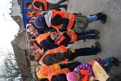 Eine Gruppe von Menschen in reflektierenden orangefarbenen Westen steht draußen, möglicherweise auf einem Bauernhof, einige tragen Mützen und Stiefel. Einige Kinder sind anwesend, eines trägt eine rosa Mütze. Rechts ist ein Holztisch zu sehen.