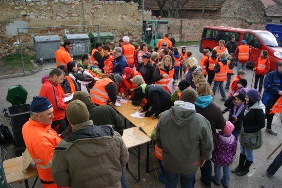 Eine Gruppe von Menschen in orangefarbenen Westen versammelt sich um einen Tisch, möglicherweise für eine Gemeinschaftsveranstaltung. Einige unterzeichnen Papiere, während andere stehen und beobachten. Ein roter Van ist in der Nähe geparkt.