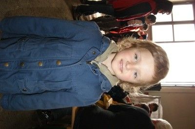 Ein junges Mädchen mit blonden Haaren, das eine blaue Jeansjacke mit Knöpfen trägt, lächelt in die Kamera in einem Raum mit anderen Personen im Hintergrund.