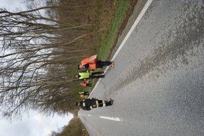 Eine Gruppe von Personen in Sicherheitswesten steht auf einer nassen Straße, möglicherweise bei einer Umfrage oder Inspektion. Bäume säumen die Straße, und der Himmel ist bewölkt.