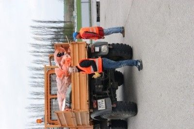 Drei Arbeiter in orangenen Westen stehen auf einem großen Lastwagen mit großen Reifen und bereiten sich möglicherweise auf eine Aufgabe auf einer asphaltierten Straße mit Bäumen im Hintergrund vor.
