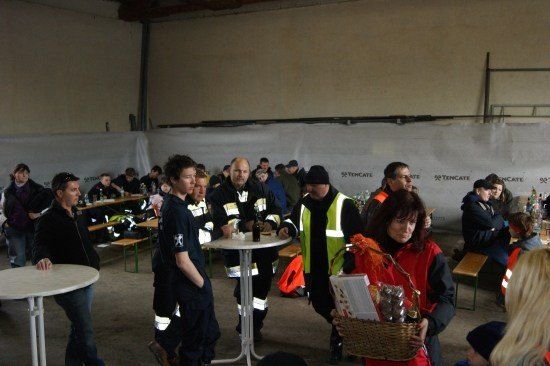 Eine Gruppe von Menschen in einem großen Raum, einige in Uniformen, andere in Freizeitkleidung. Eine Frau hält einen Korb und geht auf einen Tisch zu.