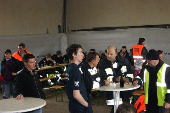Eine Gruppe von Menschen in einem Raum mit Tischen und Bänken, einige tragen Sicherheitswesten und andere Jacken. Ein Mann in einem schwarzen Jackett steht und spricht mit einem Mann in einem schwarzen Shirt.