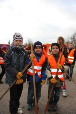 Drei Kinder in orangefarbenen Westen und Handschuhen, die Holzstäbe halten, stehen zusammen in einer Gruppe, mit anderen Personen und einem verschwommenen Hintergrund.
